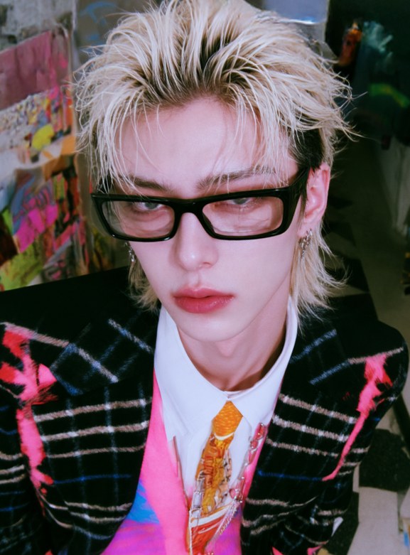 VESPER with bleached blond hair, black glasses, a check jacket and a pink shirt